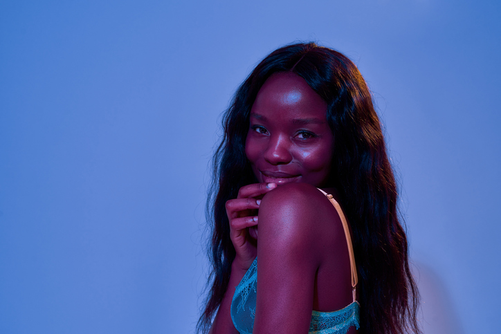 Portrait of cute african american young woman with long hair smiling at camera, looking shy while posing in lace bra isolated in neon uv purple blue light on international fetish day