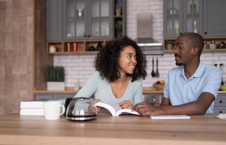 Young African American couple reading a book and learning, sitting in the kitchen, taking notes and discussing