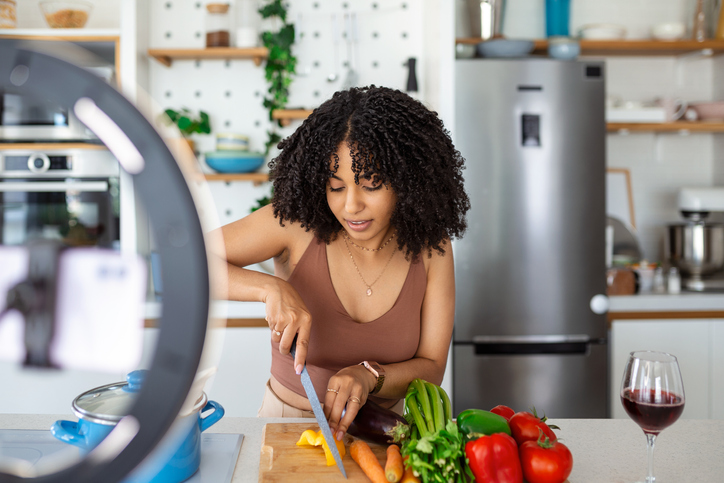 Cheerful food blogger wearing apron standing at table in modern kitchen cutting vegetables, shooting this process on camera