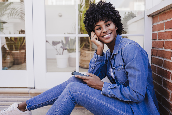 Happy young woman with smart phone sitting in doorway thinking of how to be happy