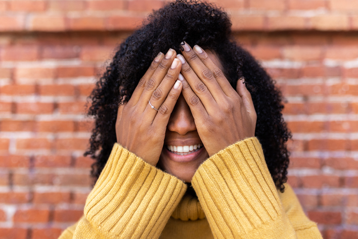 Smiling African American woman with curly hair covering her eyes with hands