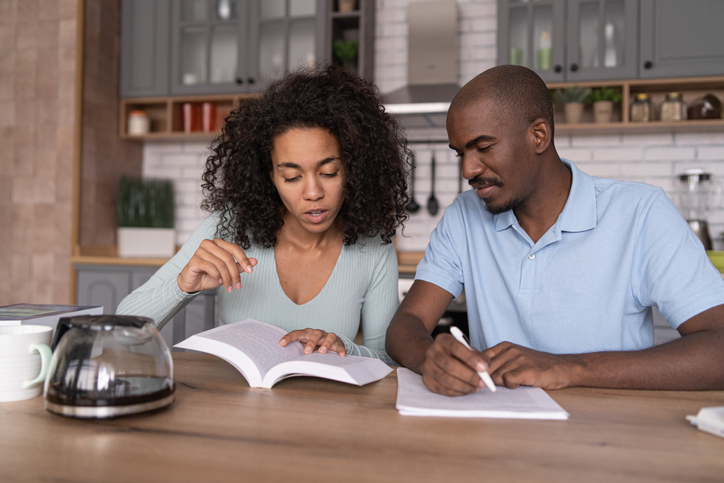 Young African American couple reading a book and learning, sitting in the kitchen, taking notes and discussing