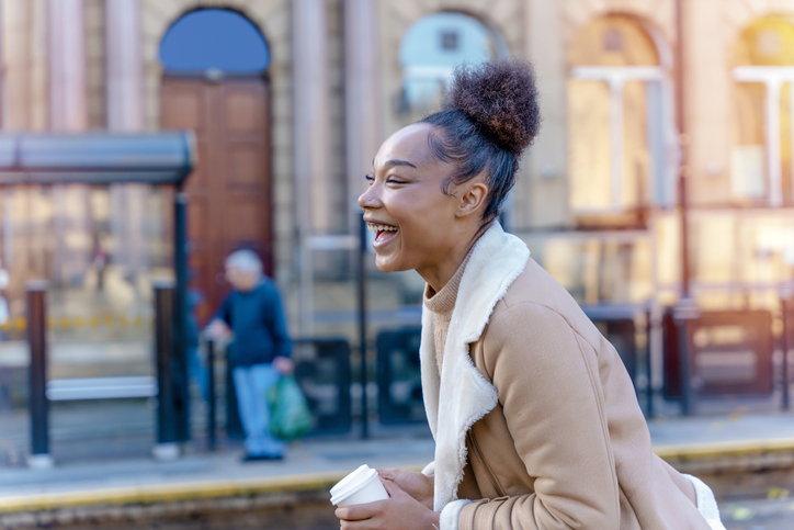 A cheerful mixed-ethnicity woman holding a cup of coffee. A smiling curly brunette lady in a sweater waiting for a tram. portrait of a single woman