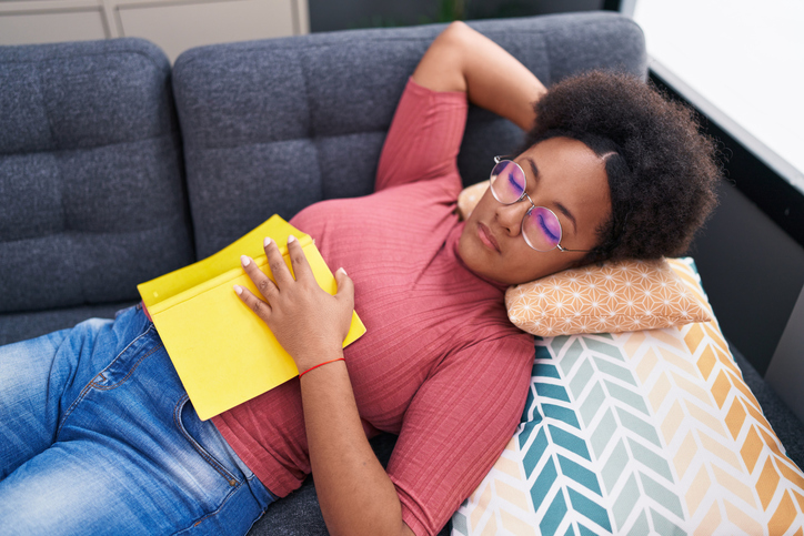 African american woman holding book lying on sofa sleeping at home