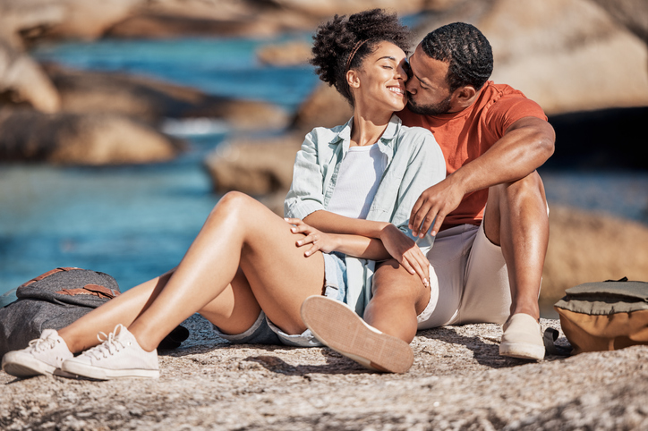 Valentine's Day getaways are for lovers. Love, kiss and happy couple on a rock at the beach relaxing, bonding and embracing while on vacation. Happiness, smile and young man and woman by the ocean on a romantic, calm and zen seaside holiday