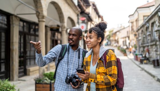 Young african couple of tourists with smartphone looking for destination and taking pictures on camera while walking on the street in an old town in Europe