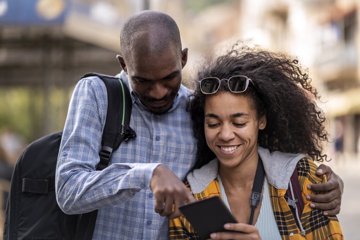 Young african couple of tourists with smartphone looking for destination and taking pictures on camera while walking on the street in an old town in Europe