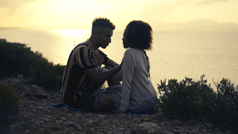 African ethnicity loving couple sitting and embracing on a cliff. Romantic sunset