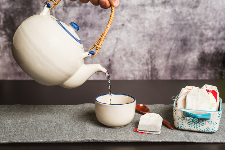 A hand holding a teapot while pouring hot water to prepare the tea, along with several sachets with different types of tea, close-up.