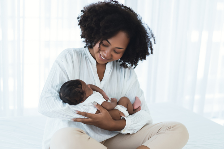 Beautiful African American young adult mother sitting on bed and holding newborn baby child in her arms, looking down at him smiling. Happy Black family lovely, nursery breastfeeding mother’s day.
