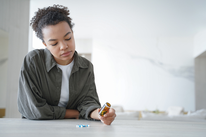 Pensive young woman holds bottle with medicines doubt whether to take pills. Healthcare. Copy space