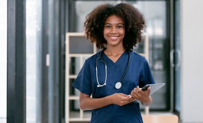 Portrait of confident, happy female nurse or doctor with stethoscope and tablet standing in hospital hallway, smiling to camera.