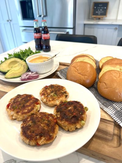 Jerk salmon burger patties on a plate, next to a plate of fresh toppings like sliced avocado and red onions, burger buns, and two bottles of Coca Cola.