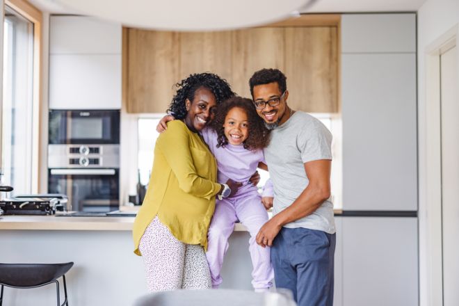 Happy Black Parents Hugging Their Smiling Black Daughter While Standing Next To The Kitchen Counter Of A Beautiful Modern Apartment