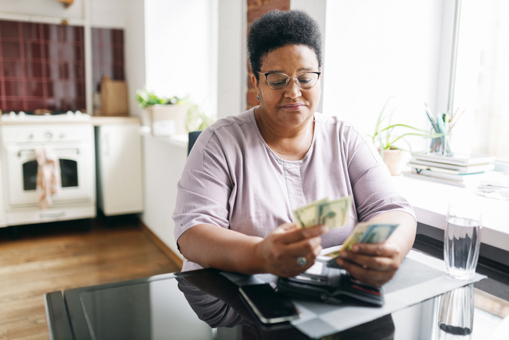 Sad frustrated plus size retired african american woman holding dollars in hands, counting her money, upset with low pension sitting at kitchen table next to window. Retired people and problems