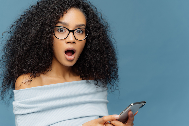 Close up portrait of stupefied young woman with Afro hairstyle, keeps mouth widely opened, holds smartphone, receives shocking message, wears blue sweater, shows bare shoulders, poses indoor
