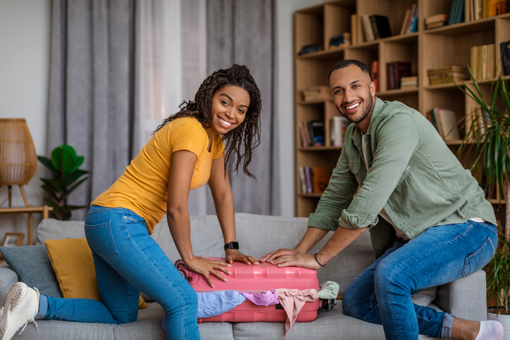 Happy black couple preparing for their trip, packing things together and trying to close suitcase at home