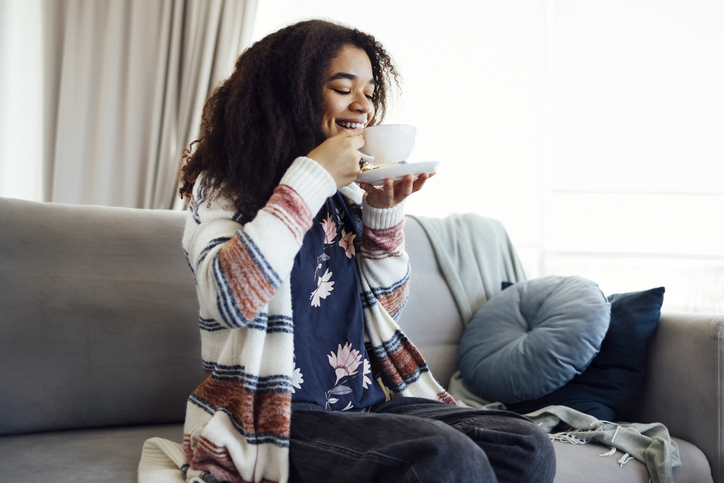 Cheerful african american girl relaxing on couch with cup of coffee, smiling at camera