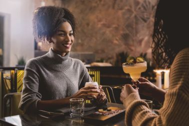 Joyful young african american woman talking to her friend - two girlfriends chatting drinking cocktails at night at indoor restaurant