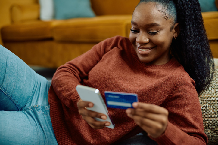 Happy black woman woman using app on mobile phone while checking her bank account at home.