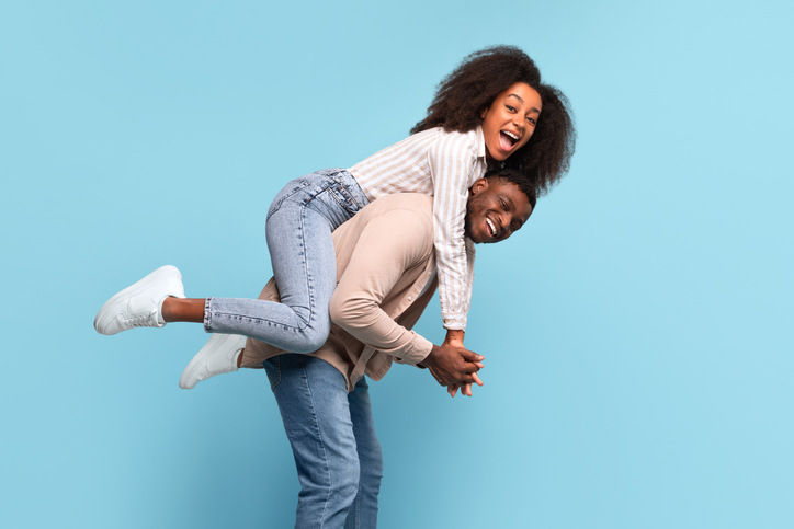 Black man giving joyful piggyback ride to woman on blue backdrop