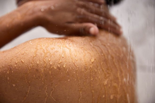 African-American woman standing side view in running shower with left arm touching neck and left shoulder detailed with water droplets