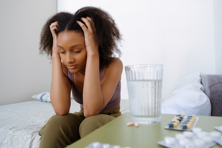 A young dark-skinned lady with a glass of water in her hands, takes a pill on the bed, feels bad at home, in the bedroom. A woman who uses a medical drug or painkiller, suffering from headaches, migraines, depression. Medical treatment. Hangover syndrome.