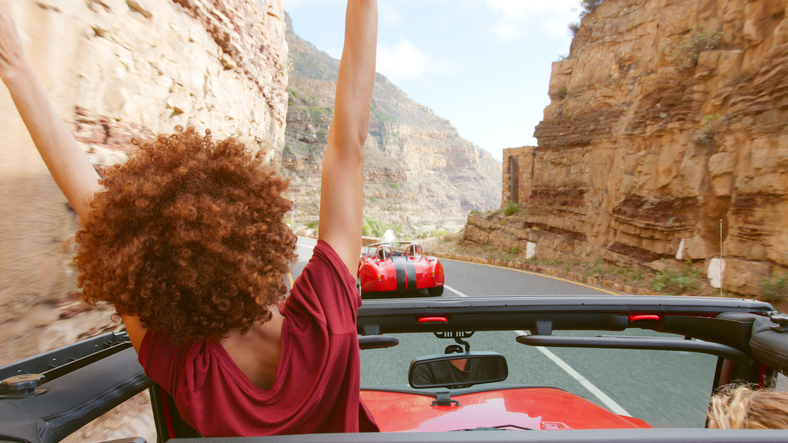 Young Female Passenger Standing Up In Open Top Hire Car Dring Along Road On Summer Vacation