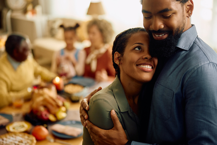 Affectionate black couple embracing while celebrating Thanksgiving with their family at home.