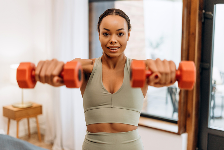 Happy athletic African American woman in sports clothes having training with weights at home