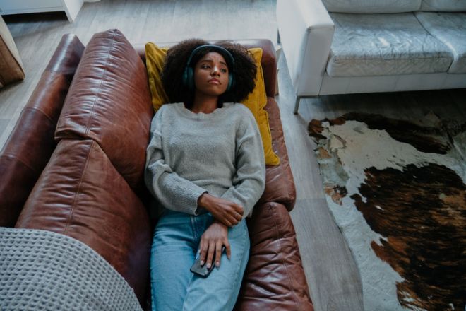 melancholic mixed race female wearing headphones listening to sad moving while holding smartphone lying on leather sofa in modern lounge