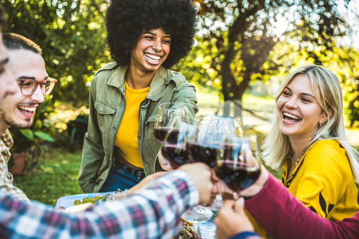Happy friends having bbq dinner party in restaurant garden - Group of young people drinking red wine and eating meat sitting at lunch table - Food and beverage lifestyle concept