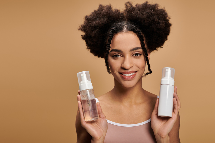 Young beautiful woman holding two skincare bottles with a smile against a warm background