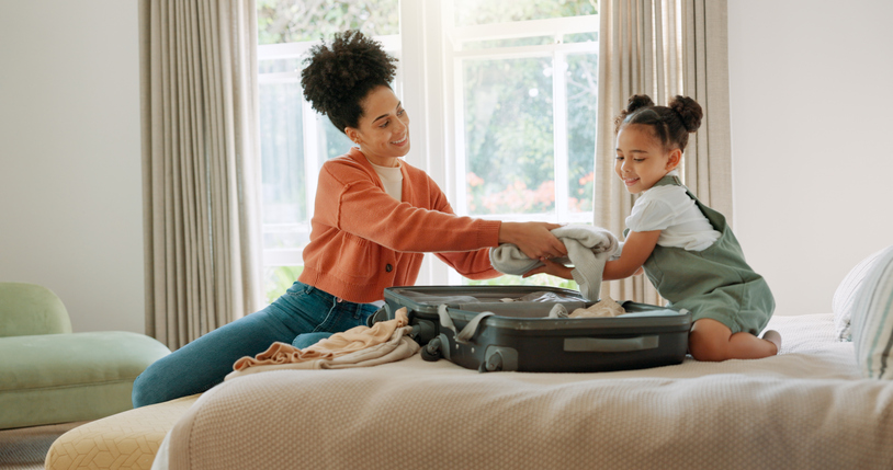 Mother, child and packing suitcase in home bedroom together for travel trip and ready for family vacation. Happy African woman or parent with girl kid on hotel bed with clothes or luggage for holiday