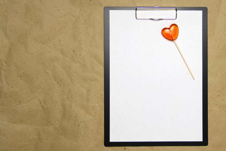 The tablet with a clip for the clip of paper with a white sheet a4 lies against the background of craft brown crumpled paper. On top lies a caramel lollipop of orange red in the shape of a heart. Place for text, copy space and layout for design