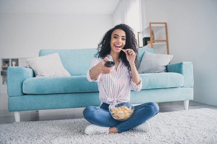 Full size photo of nice young girl sit floor watch tv remote eat chips wear striped shirt white interior apartment indoors