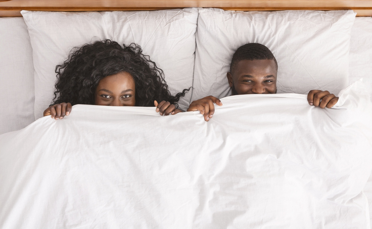 Playful black couple hiding under white blanket in bed