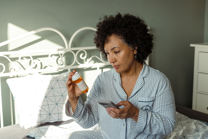 Mature woman in pajamas examining medication bottle while referring to phone