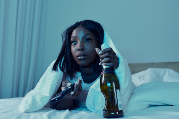 Young black woman watching TV with a bottle of wine on a bed at night.