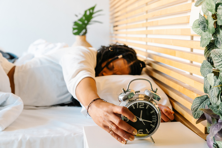 Drowsy female relaxing in bedroom, stretching to silence morning alarm, soft smile emerging while waking up