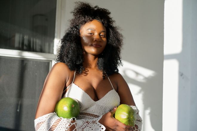 Woman in white crochet top holding two large green fruits, standing in sunlight. Miami, Florida, USA