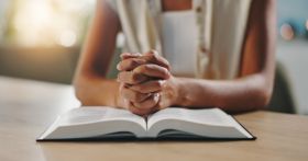 Hands, praying and bible in home with religion, study and hope for change, spiritual or reflection in morning. Person, book and scripture with prayer, worship or learning for Christian faith in house