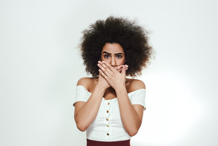 Never say too much. Young afro american woman with curly hair is covering mouth with hands and looking at camera while standing against grey background