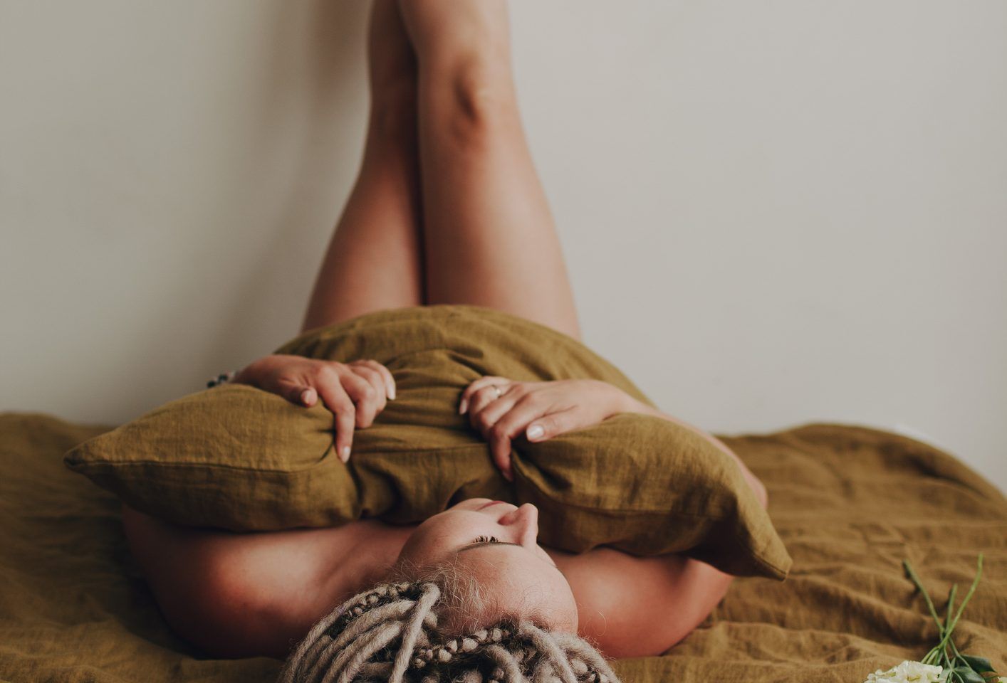 A half-naked girl with dreadlocks is lying on a linen sheet, her bare legs are raised up and crossed, leaning against a white wall, her body is covered with a pillow