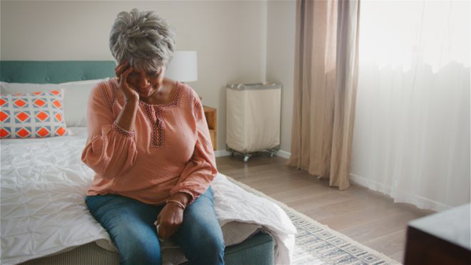 Unhappy Senior Woman With Head In Hands Suffering With Depression Sitting In Bedroom At Home
