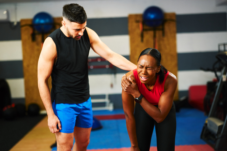 Sportswoman feeling strong shoulder pain during training session with personal trainer assisting her