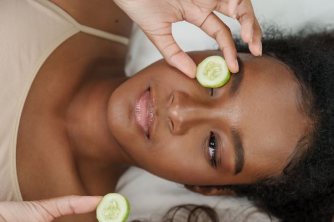 multiracial teenage girl lying bed holding cucumber slices over eyes showing skincare routine natural beauty home spa self care glow fresh skin youthful health relaxing moment