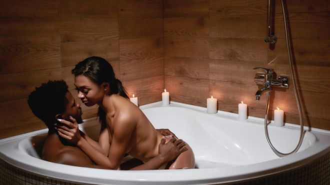 Intimate moment. Young sensual naked african couple of lovers kissing while taking foamy bath with candles together