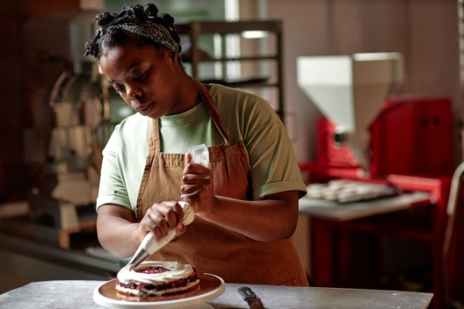 Black Female Pastry Chef Icing Cake with Cream Using Piping Bag in Confectionery Store Kitchen