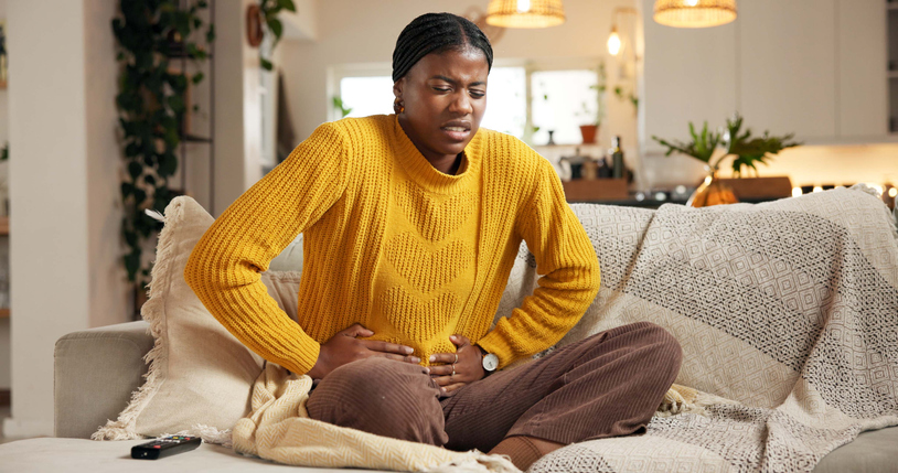 Stomach ache, couch or black woman with pain in living room, constipation or menstruation cycle for sore belly. Upset, period cramps or person with abdominal problem, digestion trouble or pms in home